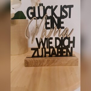 Pu&ograve; includere: Un cartello in legno nero con il testo "Gl&uuml;ck ist eine Mama wie dich zu haben" che si traduce in "La felicit&agrave; &egrave; avere una mamma come te". Il cartello &egrave; su una base in legno.