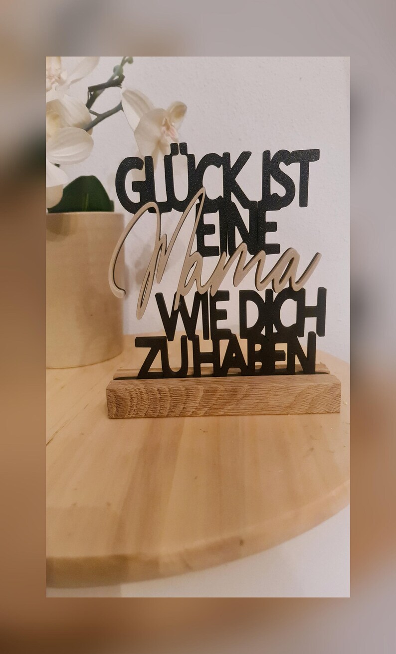 Pu&ograve; includere: Un cartello in legno nero con il testo "Gl&uuml;ck ist eine Mama wie dich zu haben" che si traduce in "La felicit&agrave; &egrave; avere una mamma come te". Il cartello &egrave; su una base in legno.