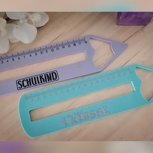 May include: Two pencil-shaped rulers, one lavender and one teal, with measurement markings and cut-out shapes. The lavender ruler has the word "SCHULKIND" printed on it. The teal ruler has the text "1. Klasse" printed on it.