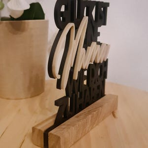 Pu&ograve; includere: Un cartello in legno nero con il testo "Gl&uuml;ck ist eine Mama wie du, wir haben zu haben" in lettere bianche. Il cartello &egrave; su un supporto in legno.