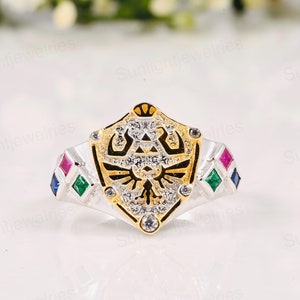 May include: A silver ring with a gold shield design featuring a stylized bird and a triangle. The shield is encrusted with clear stones and the ring is accented with blue, green, and red stones.