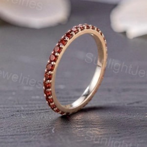 Puede incluir: Un anillo de metal fino con una banda continua de pequeñas piedras preciosas redondas de color rojo oscuro engastadas a lo largo de la circunferencia exterior. La banda tiene un acabado metálico pulido y el texto SunlightJewelries es visible.