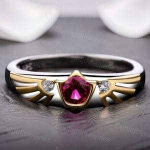 May include: A silver-toned ring featuring a central pentagonal setting with a vibrant pink gemstone. The band is accented with gold-toned wing-like motifs on either side, each embellished with a small, clear, round-cut white gemstone.