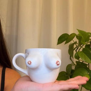 Peut inclure: Une tasse en céramique blanche présentant un design sculpté en trois dimensions représentant deux seins sur le devant. La tasse a une finition lisse et brillante avec une anse incurvée sur le côté gauche et deux petits détails texturés roses.