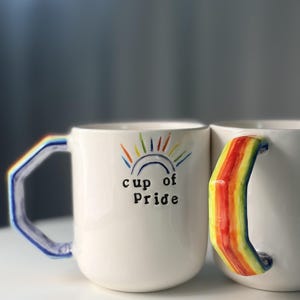Könnte beinhalten: Zwei weiße Keramiktassen mit handbemalten Regenbogenhenkeln. Auf der Vorderseite einer Tasse befindet sich ein kleiner, bunt bemalter Regenbogenbogen mit Sonnenstrahlen darüber und dem schwarzen Text "cup of Pride" darunter.