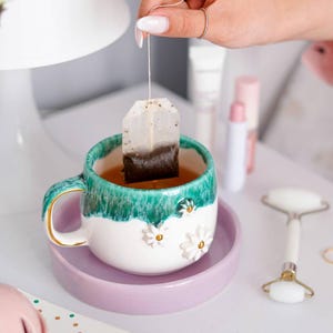 Op de afbeelding: Een keramische mok met een witte basis en een blauwgroene druipglazuur rand, voorzien van driedimensionale witte madeliefjes met gele harten. De mok staat op een lichtpaarse schotel en bevat een theezakje in een amberkleurige vloeistof.