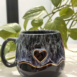 May include: A ceramic mug featuring a glossy black base and a textured, drippy glaze in shades of blue and white on the upper portion. A raised, dark heart shape is centered on the front, outlined by a thin, metallic gold wavy line.