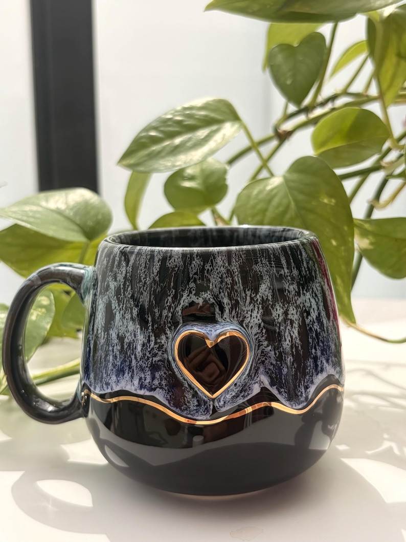 May include: A ceramic mug featuring a glossy black base and a textured, drippy glaze in shades of blue and white on the upper portion. A raised, dark heart shape is centered on the front, outlined by a thin, metallic gold wavy line.