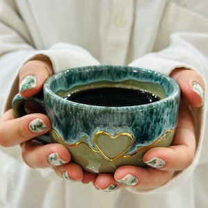 Könnte beinhalten: Eine Keramik-Kaffeetasse mit herzförmigem Design und goldenem Rand. Die Tasse hat eine türkis-grüne Glasur und ist mit dunkler Flüssigkeit gefüllt. Die Tasse wird von einer Person gehalten, die ein weißes Hemd trägt.