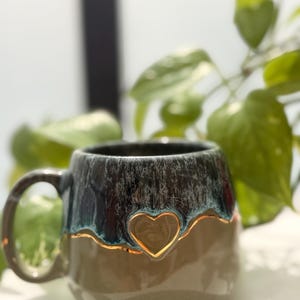 May include: A ceramic mug featuring a two-tone glaze with a dark, mottled blue-grey top section and a smooth, light beige bottom. A raised heart shape with a metallic gold-colored outline is centered on the front where the two colors meet.
