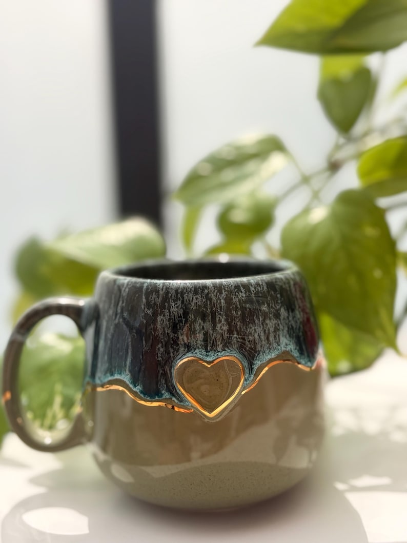 May include: A ceramic mug featuring a two-tone glaze with a dark, mottled blue-grey top section and a smooth, light beige bottom. A raised heart shape with a metallic gold-colored outline is centered on the front where the two colors meet.