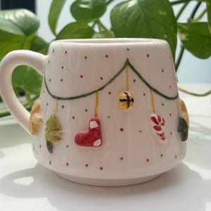 May include: A cream-colored ceramic mug featuring a hand-painted design of green garland draped across the surface. The mug is decorated with small red and green dots and raised, three-dimensional ornaments including a stocking and candy cane.