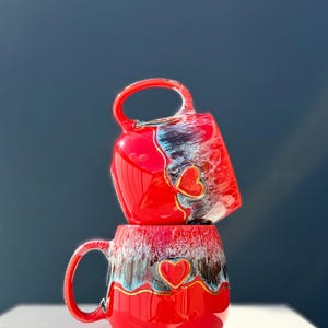 May include: Two stacked ceramic mugs featuring a vibrant red glaze with a decorative drip pattern in shades of white, blue, and black. Each mug is adorned with a raised, gold-outlined red heart emblem centered on the front surface.