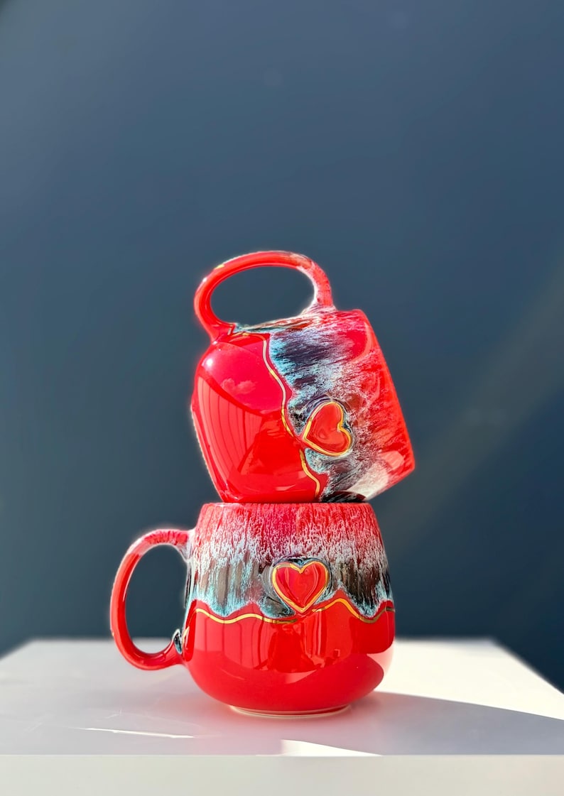 May include: Two stacked ceramic mugs featuring a vibrant red glaze with a decorative drip pattern in shades of white, blue, and black. Each mug is adorned with a raised, gold-outlined red heart emblem centered on the front surface.