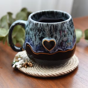 May include: A ceramic mug featuring a glossy black base and a textured, drippy glaze in shades of blue and white. A small, raised heart shape with a gold-colored outline is centered on the front, separated from the black base by a thin gold line.