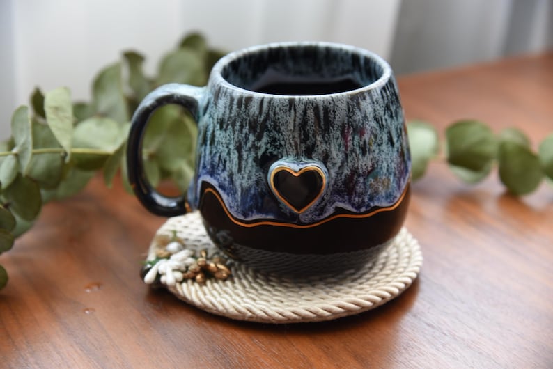 May include: A ceramic mug featuring a glossy black base and a textured, drippy glaze in shades of blue and white. A small, raised heart shape with a gold-colored outline is centered on the front, separated from the black base by a thin gold line.