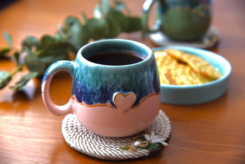 May include: A ceramic mug with a rounded shape featuring a two-tone glaze of deep blue and green at the top and soft pink at the bottom. A small, raised pink heart emblem is centered on the front, accented with a thin gold-colored trim line.