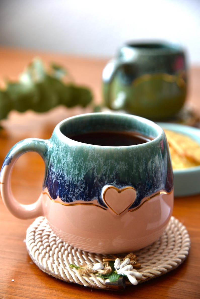 May include: A round ceramic mug featuring a two-tone glaze with a soft pink base and a dark blue and green top. A small, raised heart shape is centered on the front, outlined in gold. The mug has a curved handle and a smooth, glossy finish.