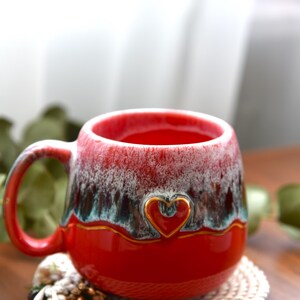 May include: A ceramic mug with a glossy red base and a textured, drippy glaze in shades of white, dark brown, and teal near the rim. The mug features a small, embossed heart outline centered on the front, highlighted with a thin gold-colored line.