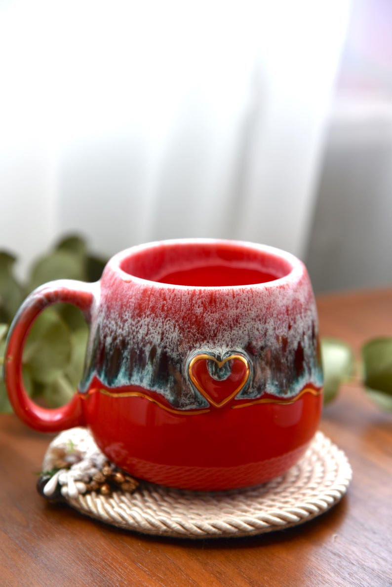 May include: A ceramic mug with a glossy red base and a textured, drippy glaze in shades of white, dark brown, and teal near the rim. The mug features a small, embossed heart outline centered on the front, highlighted with a thin gold-colored line.
