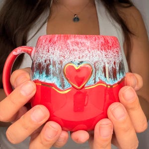 May include: A ceramic mug featuring a vibrant red base and a textured upper section with white and teal drip glaze patterns. A raised, glossy red heart emblem is centered on the front, accented by a thin, horizontal gold line that wraps around.
