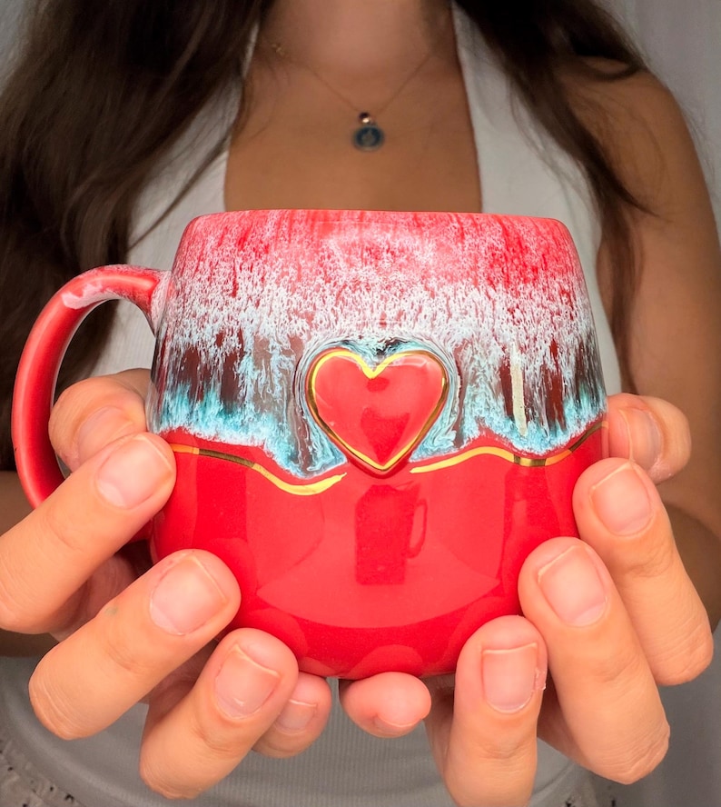 May include: A ceramic mug featuring a vibrant red base and a textured upper section with white and teal drip glaze patterns. A raised, glossy red heart emblem is centered on the front, accented by a thin, horizontal gold line that wraps around.