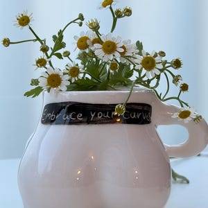 Könnte beinhalten: Ein weißer Keramikbecher in Form eines menschlichen Torsos mit einem Henkel auf der rechten Seite. Ein schwarzer horizontaler Pinselstrich auf der Vorderseite trägt den weißen Text "Embrace your curves". Der Becher ist mit weißen Margeriten gefüllt.