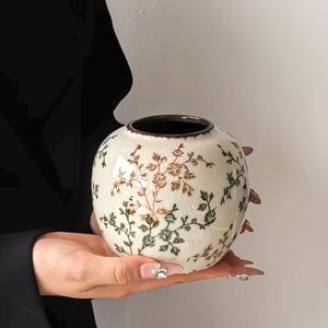 Peut inclure: Un vase en céramique de forme ronde, de couleur crème, doté d'une finition émaillée craquelée. La surface est ornée d'un motif répétitif de petites vignes florales délicates dans des tons de vert et de brun. Le vase possède un bord brun foncé.