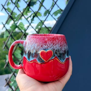 Può includere: Una tazza in ceramica rotonda con una base rosso vivace e uno smalto lucido che sfuma in un motivo a goccia bianco e blu testurizzato sulla parte superiore. Un cuore rosso tridimensionale in rilievo con un bordo dorato è centrato sul davanti.