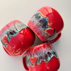 May include: Three ceramic mugs with a glossy red finish, featuring a textured blue and white drip glaze pattern along the upper rim. Each mug is accented with a small, raised red heart shape outlined in gold, positioned on the side.
