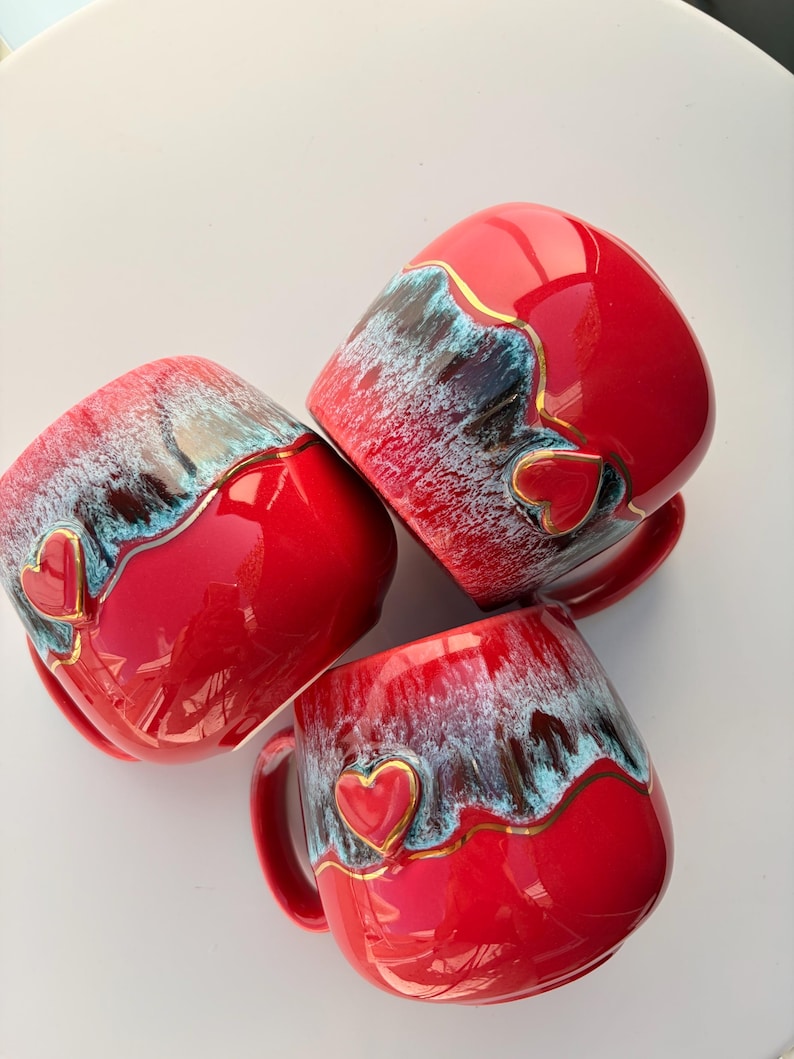 May include: Three ceramic mugs with a glossy red finish, featuring a textured blue and white drip glaze pattern along the upper rim. Each mug is accented with a small, raised red heart shape outlined in gold, positioned on the side.