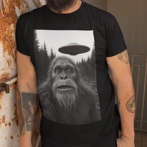 May include: Black t-shirt featuring a black and white image of Bigfoot with a UFO hovering above. The graphic tee has a forest background and a surprised expression on the Bigfoot's face. The shirt is worn by a person.
