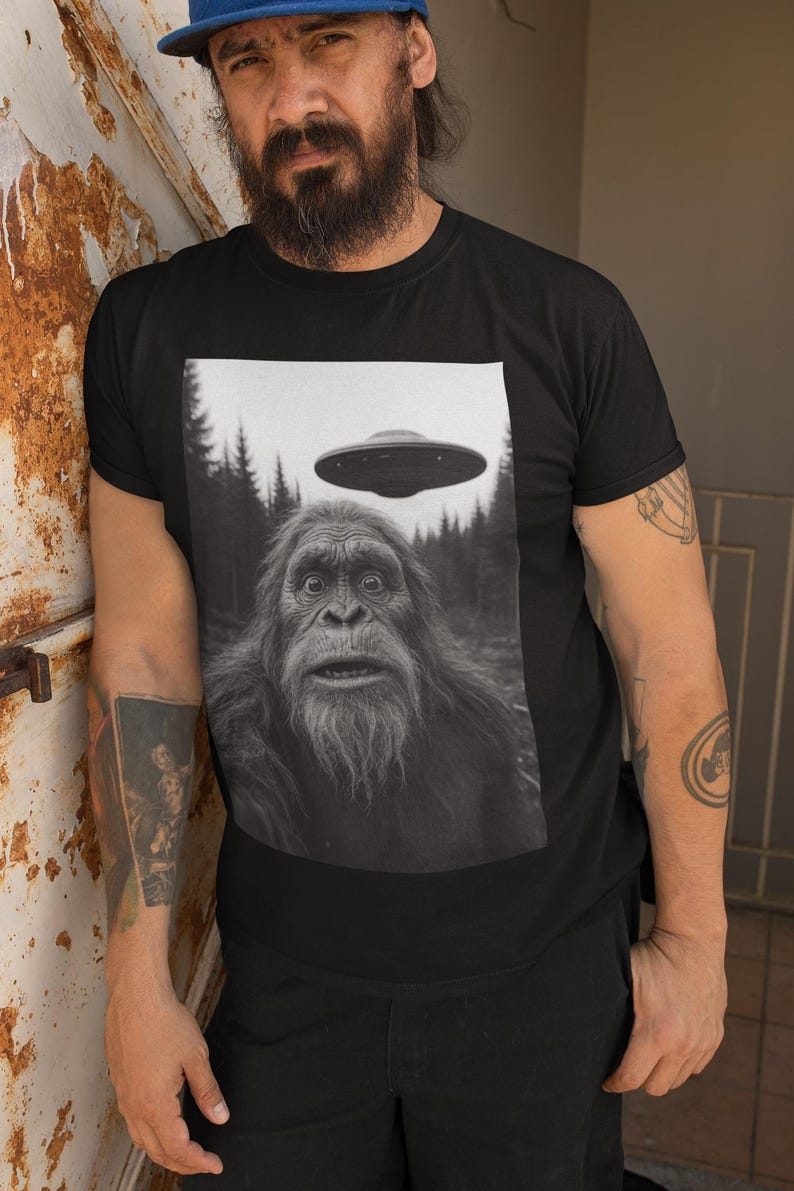 May include: Black t-shirt featuring a black and white image of Bigfoot with a UFO hovering above. The graphic tee has a forest background and a surprised expression on the Bigfoot's face. The shirt is worn by a person.
