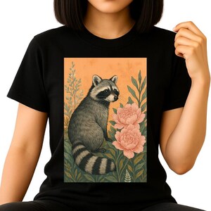 May include: Black t-shirt featuring a raccoon illustration. The raccoon is depicted in shades of gray and black, sitting amidst pink flowers and green foliage against an orange background. The design is printed on a black t-shirt.
