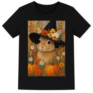 May include: Black t-shirt featuring a whimsical illustration of a bunny wearing a witch's hat adorned with flowers. The design includes pumpkins and flowers, creating a fall or Halloween theme. The artwork is set against a backdrop of wheat.