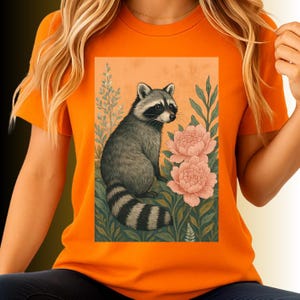 May include: Orange t-shirt featuring a raccoon illustration. The raccoon is depicted in gray and black, surrounded by pink peonies and green foliage against a peach background. The design is printed on a short-sleeved orange tee.