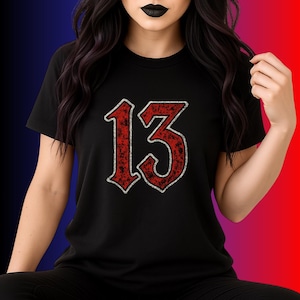 Op de afbeelding: Zwart T-shirt met het nummer "13" in een opvallend, verweerd rood lettertype met een zilveren omlijning. De afbeelding staat in het midden van het shirt. Het shirt heeft korte mouwen en een ronde hals.