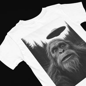 May include: White t-shirt featuring a black and white graphic of a surprised Bigfoot looking up at a UFO. The design includes a forest backdrop and a flying saucer hovering above the creature's head.