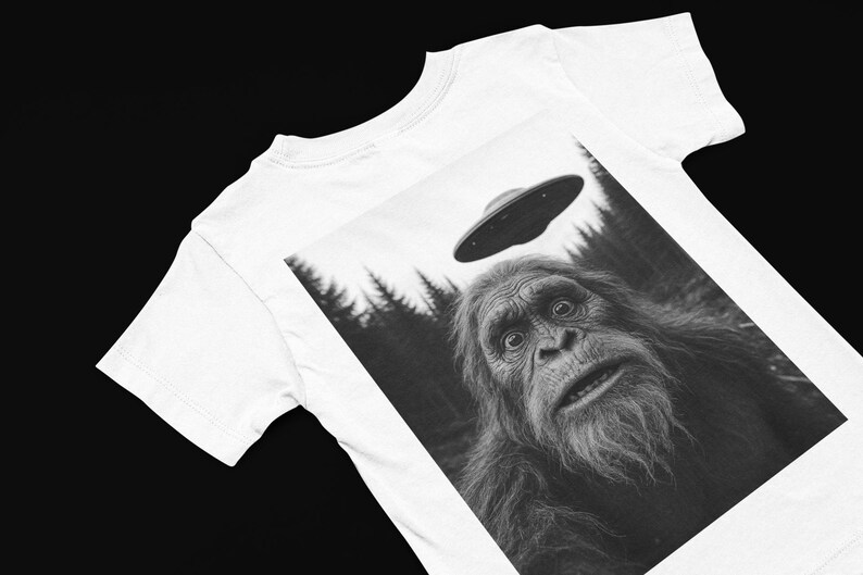 May include: White t-shirt featuring a black and white graphic of a surprised Bigfoot looking up at a UFO. The design includes a forest backdrop and a flying saucer hovering above the creature's head.