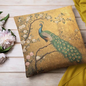 May include: Square cushion with a peacock design. The cushion has a gold background with a detailed illustration of a peacock perched on a branch, surrounded by white and pink flowers. A yellow fabric is draped nearby.