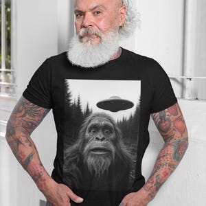 May include: Black t-shirt featuring a black and white image of Bigfoot with a UFO hovering above. The image is set against a forest backdrop. The person wearing the shirt is standing in front of a white wall.