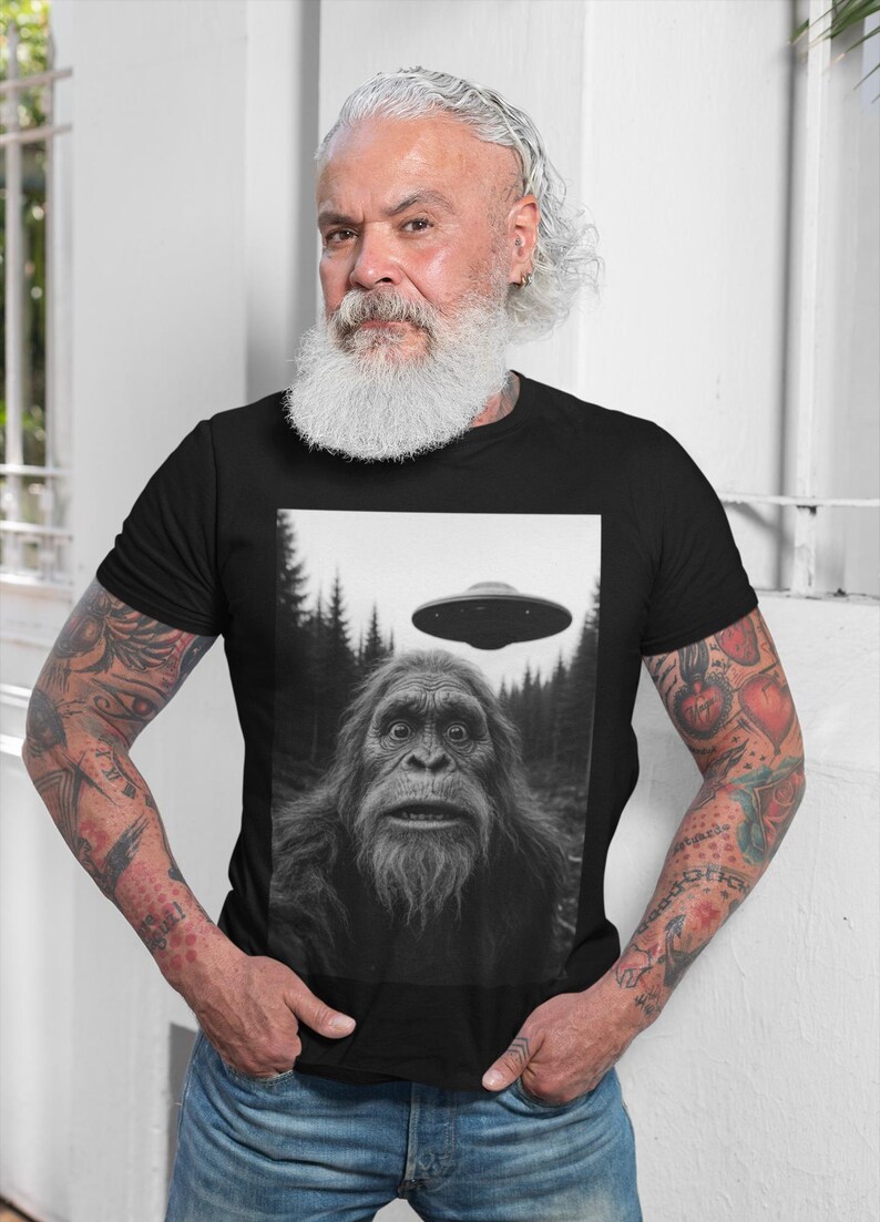May include: Black t-shirt featuring a black and white image of Bigfoot with a UFO hovering above. The image is set against a forest backdrop. The person wearing the shirt is standing in front of a white wall.