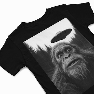 May include: Black t-shirt featuring a black and white graphic of a surprised Bigfoot looking up at a UFO. The image includes a forest background and a flying saucer above the creature's head.