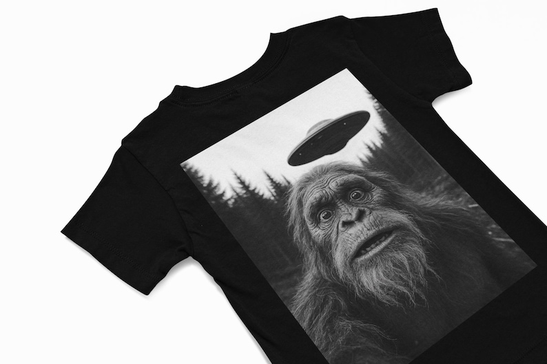 May include: Black t-shirt featuring a black and white graphic of a surprised Bigfoot looking up at a UFO. The image includes a forest background and a flying saucer above the creature's head.