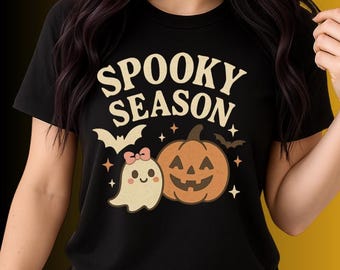 May include: Black t-shirt featuring a Halloween design. The words "Spooky Season" are above a cartoon ghost with a pink bow, a jack-o'-lantern, and a bat. The design is in cream, orange, and black.