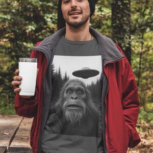May include: A dark gray t-shirt featuring a black and white graphic of a Bigfoot and a UFO. The person in the image is wearing a black beanie, a red jacket, and holding a white tumbler. The background is a forest.