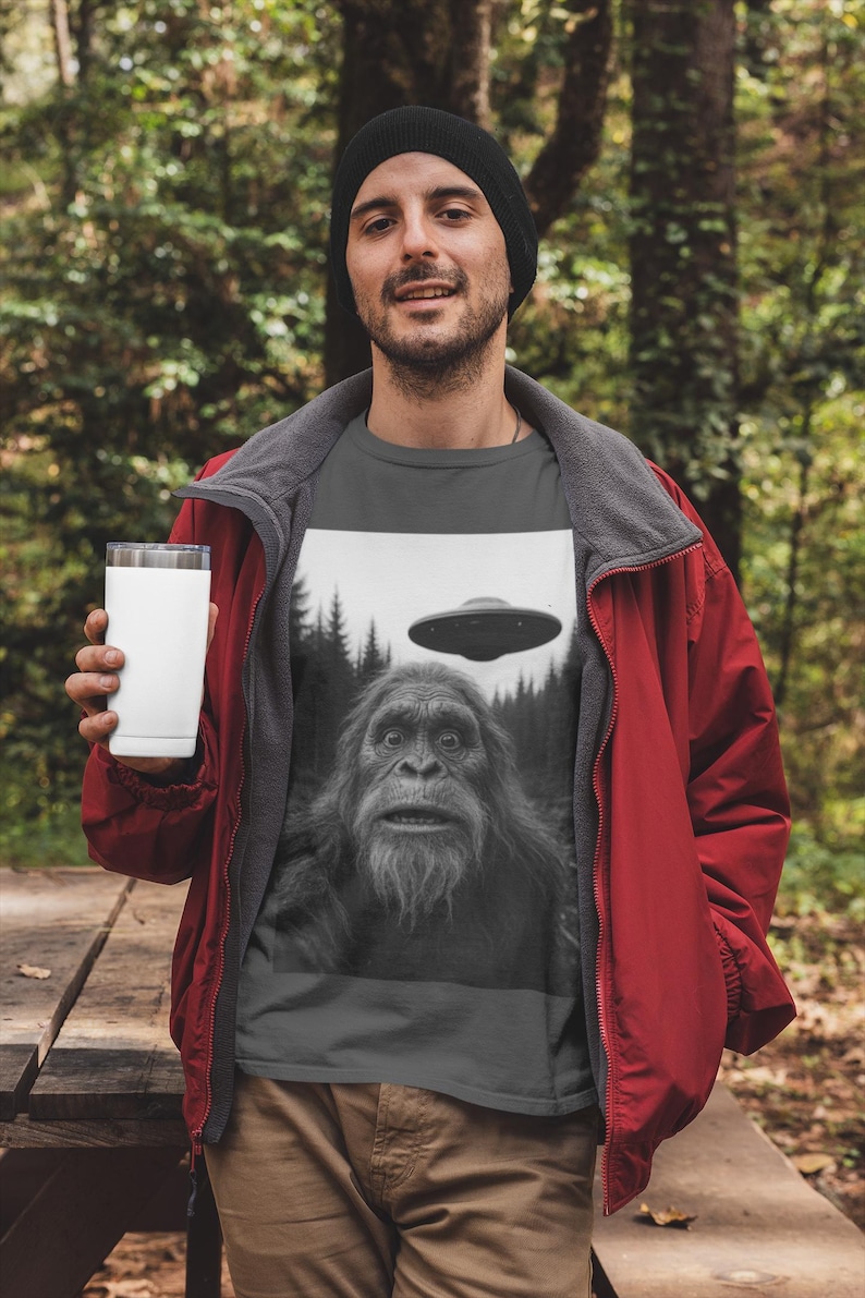 May include: A dark gray t-shirt featuring a black and white graphic of a Bigfoot and a UFO. The person in the image is wearing a black beanie, a red jacket, and holding a white tumbler. The background is a forest.