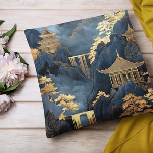 May include: Square decorative pillow with a blue and gold Asian landscape design. The pillow features pagodas, mountains, and trees in gold against a dark blue background. The pillow is on a wooden surface with flowers.