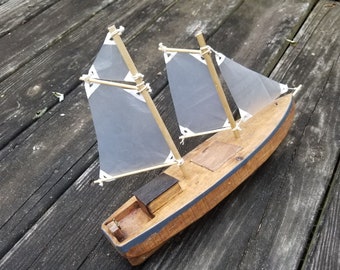 May include: A wooden model sailboat with two masts and translucent sails. The hull is made of light brown wood with a dark blue trim. The sails are a light gray color, and the boat is sitting on a weathered wooden surface.