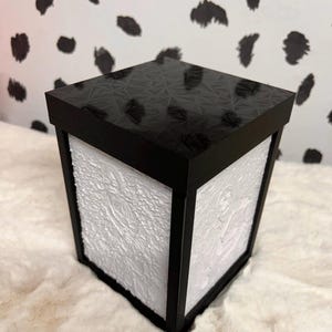 Może przedstawiać: Czarne kwadratowe pudełko z białym, teksturowanym wnętrzem. Górna część pudełka ma czarny, geometryczny wzór.